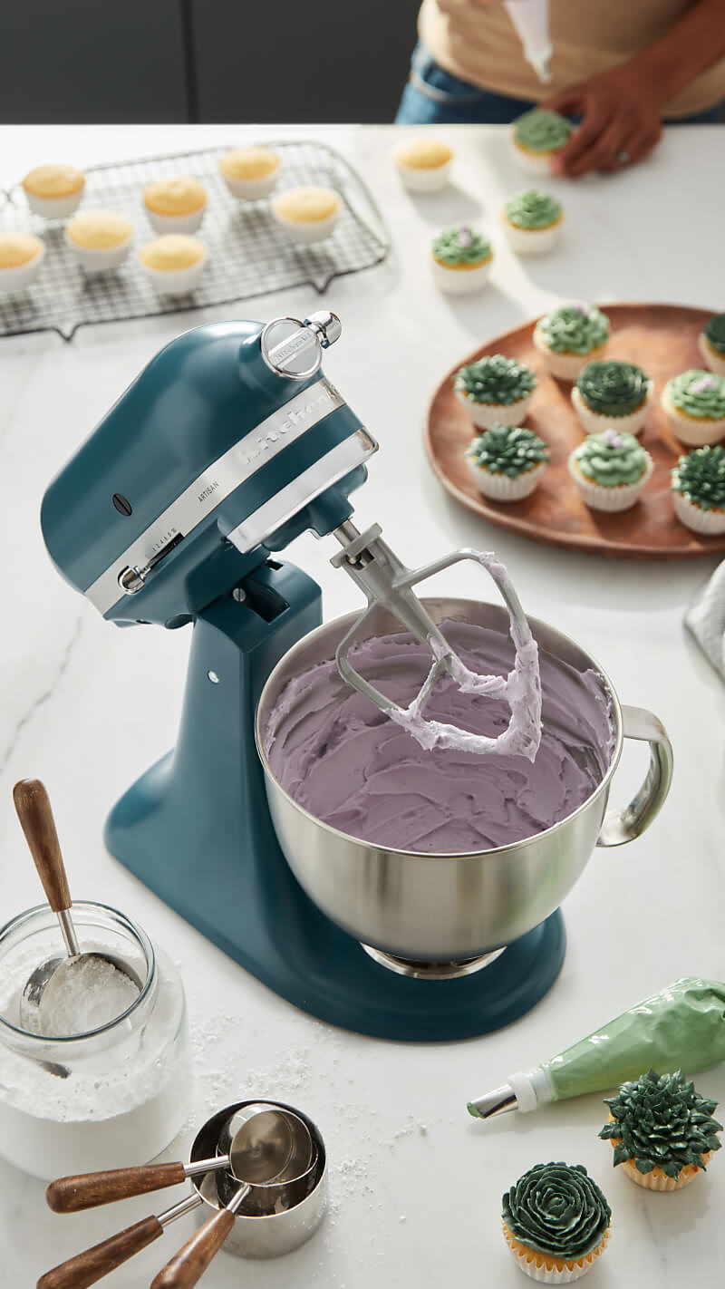 An Agave stand mixer mixing purple icing surrounded by succulent cupcakes and baking tools. 